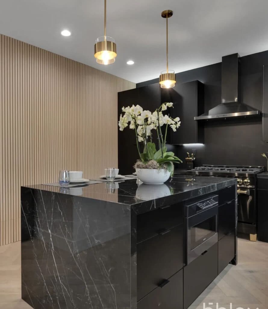 Black kitchen with a marble island, a wood-slat accent wall, brass pendants, and integrated appliances.