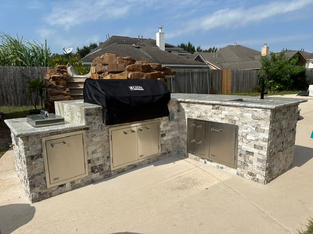 Stone-veneer outdoor kitchen island with a built-in grill, side burners, and a sink in a suburban backyard.