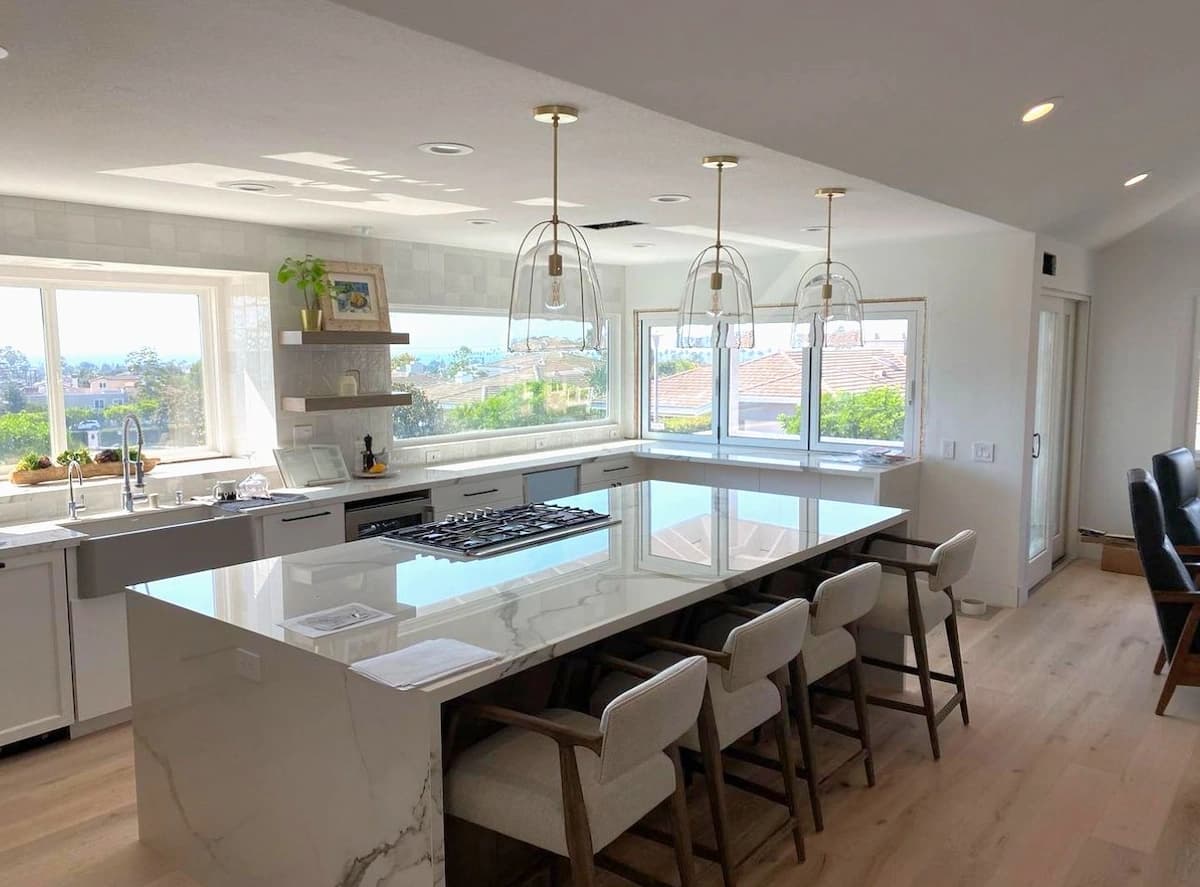 Bright white kitchen with a marble waterfall island, pendant lights, bar seating, and large view windows.