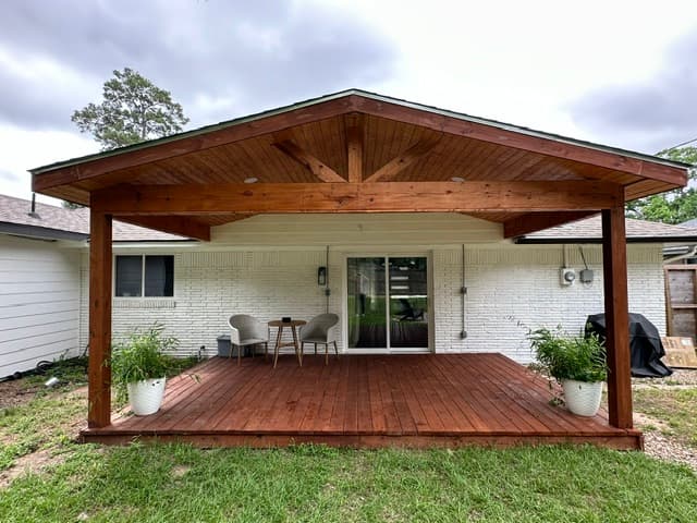 Cedar-stained gable patio cover over a wood deck on a white-brick home, with patio seating.