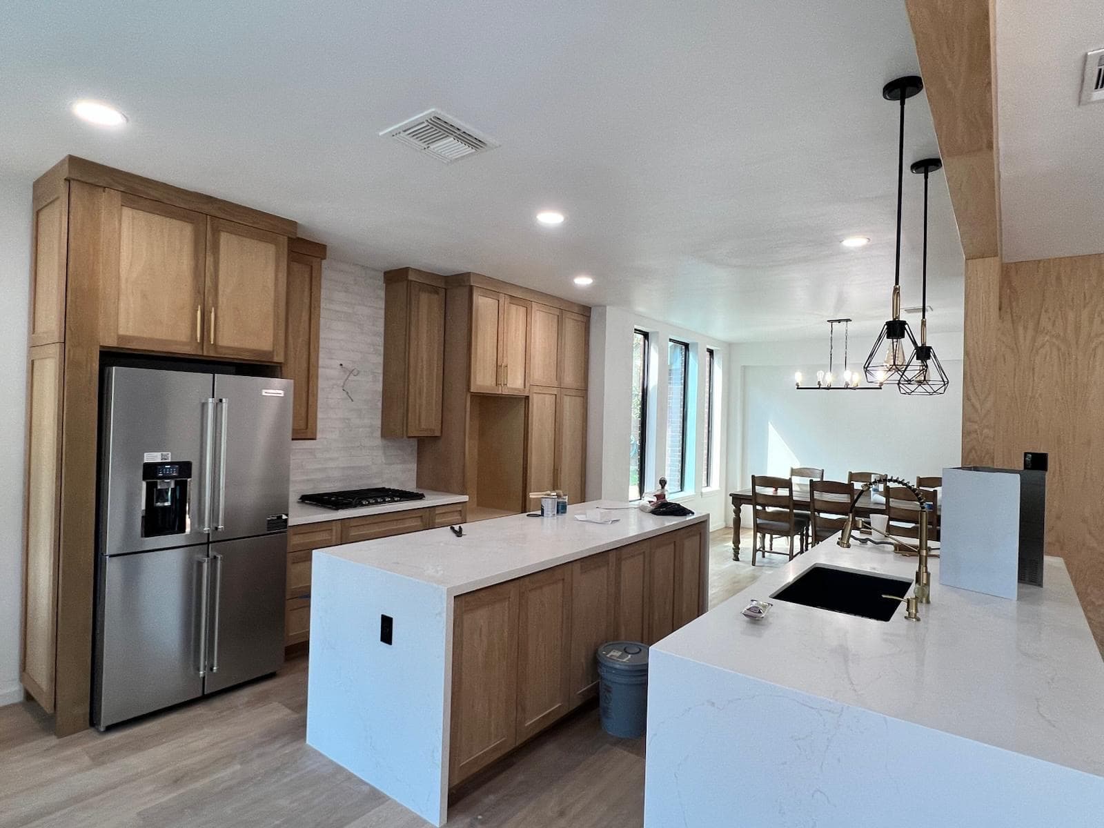 Wood-cabinet kitchen with a white quartz waterfall island, stainless refrigerator, gas cooktop, and an open dining area.