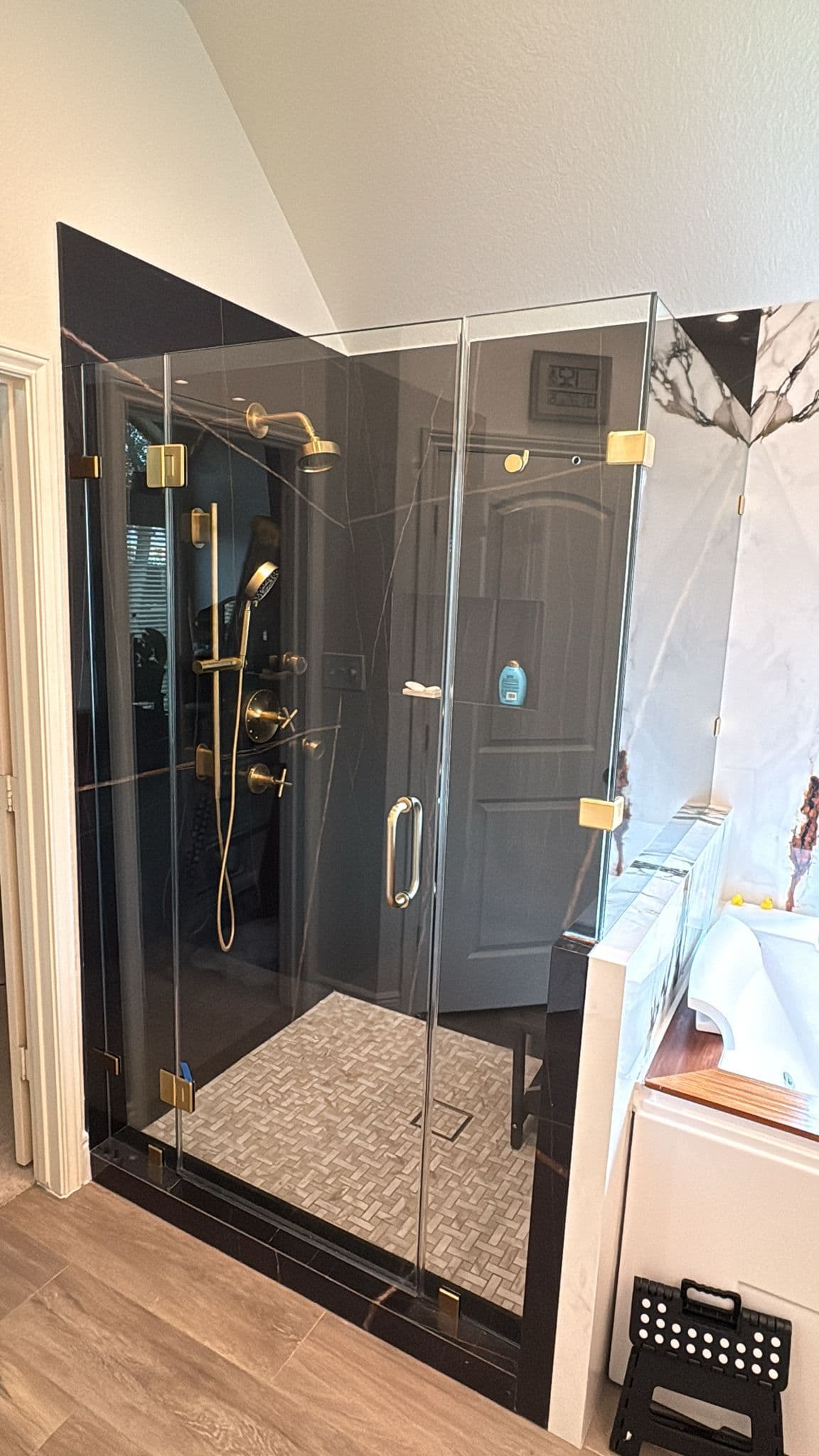 Black-marble shower enclosure with brushed-gold fixtures, a herringbone tile floor, and frameless glass doors.