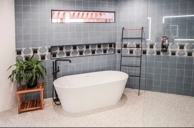 Bathroom with slate-blue square wall tile and a circular-pattern accent band, a freestanding white tub, a matte-black floor-mount tub filler, a black ladder towel rack, and a terrazzo-look floor.