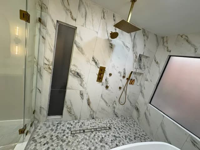 Walk-in shower with book-matched white marble walls, a brushed-brass rain head, a recessed vertical niche, and a pebble-marble floor behind glass.