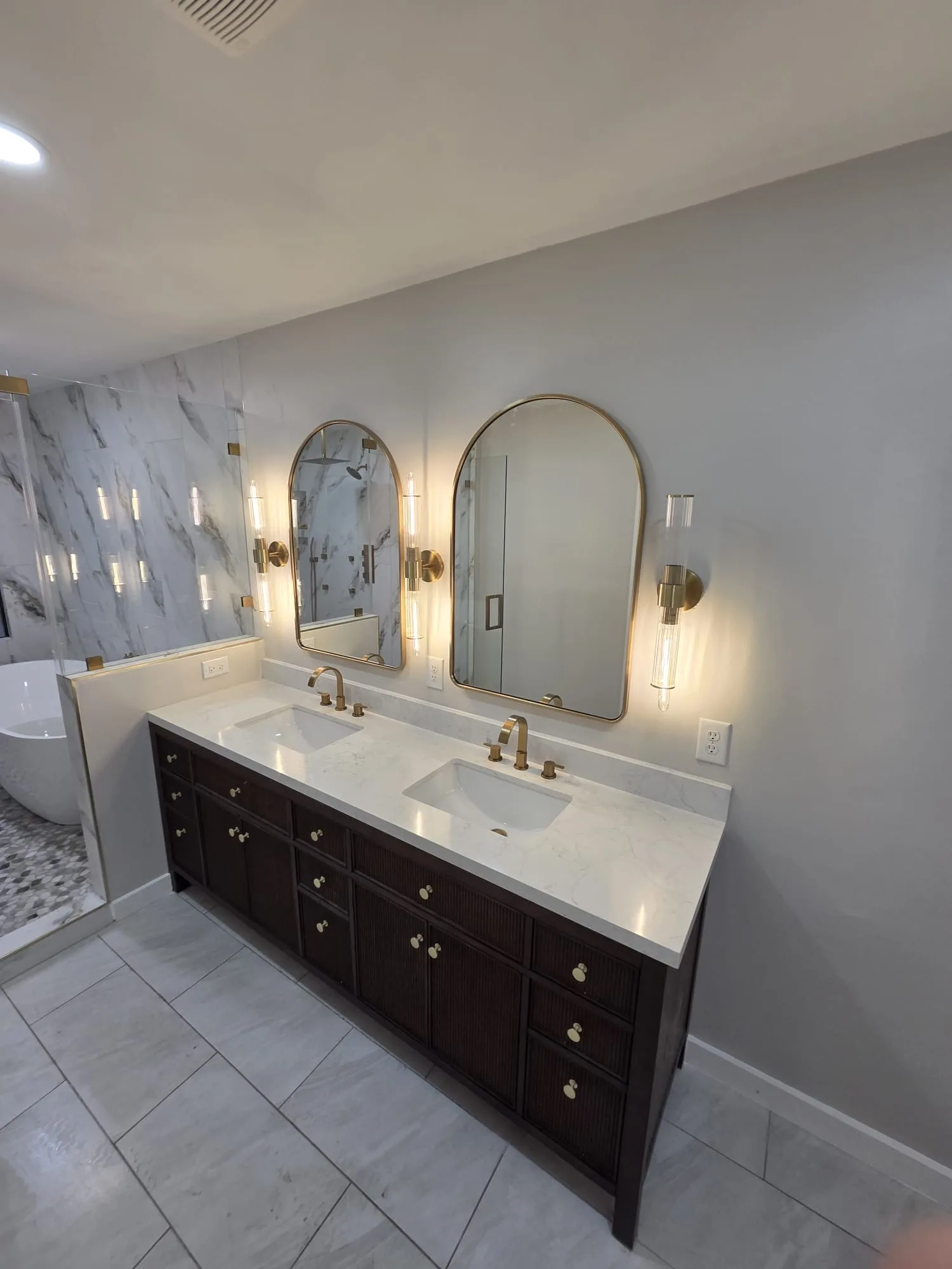 Bathroom with a dark espresso double vanity, white quartz counters, arched gold-framed mirrors, brushed-brass cylinder sconces, and white marble walls.