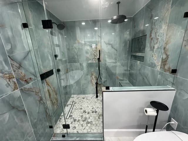 Walk-in shower clad in dramatic green-and-gold quartzite slabs, with a black rain showerhead, a hand shower, and a hexagon marble floor behind frameless glass.