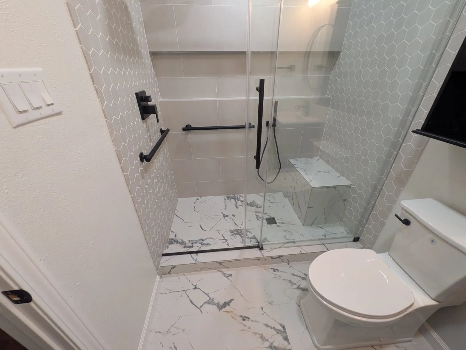 Walk-in shower with a marble-look floor, hexagon accent tile, matte-black grab bars and fixtures, a built-in marble bench, and a frameless glass enclosure.