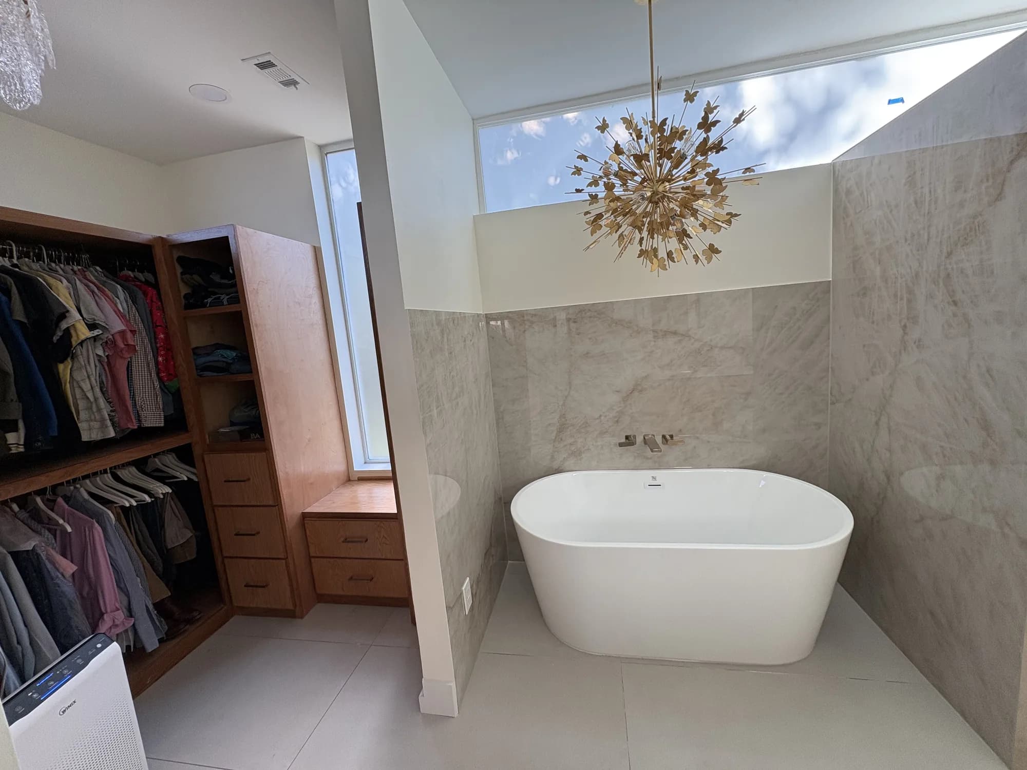 Master bathroom with a freestanding tub beneath a clerestory window, a gold sputnik-style chandelier, light marble walls, and an adjoining walk-in closet with wood shelving.