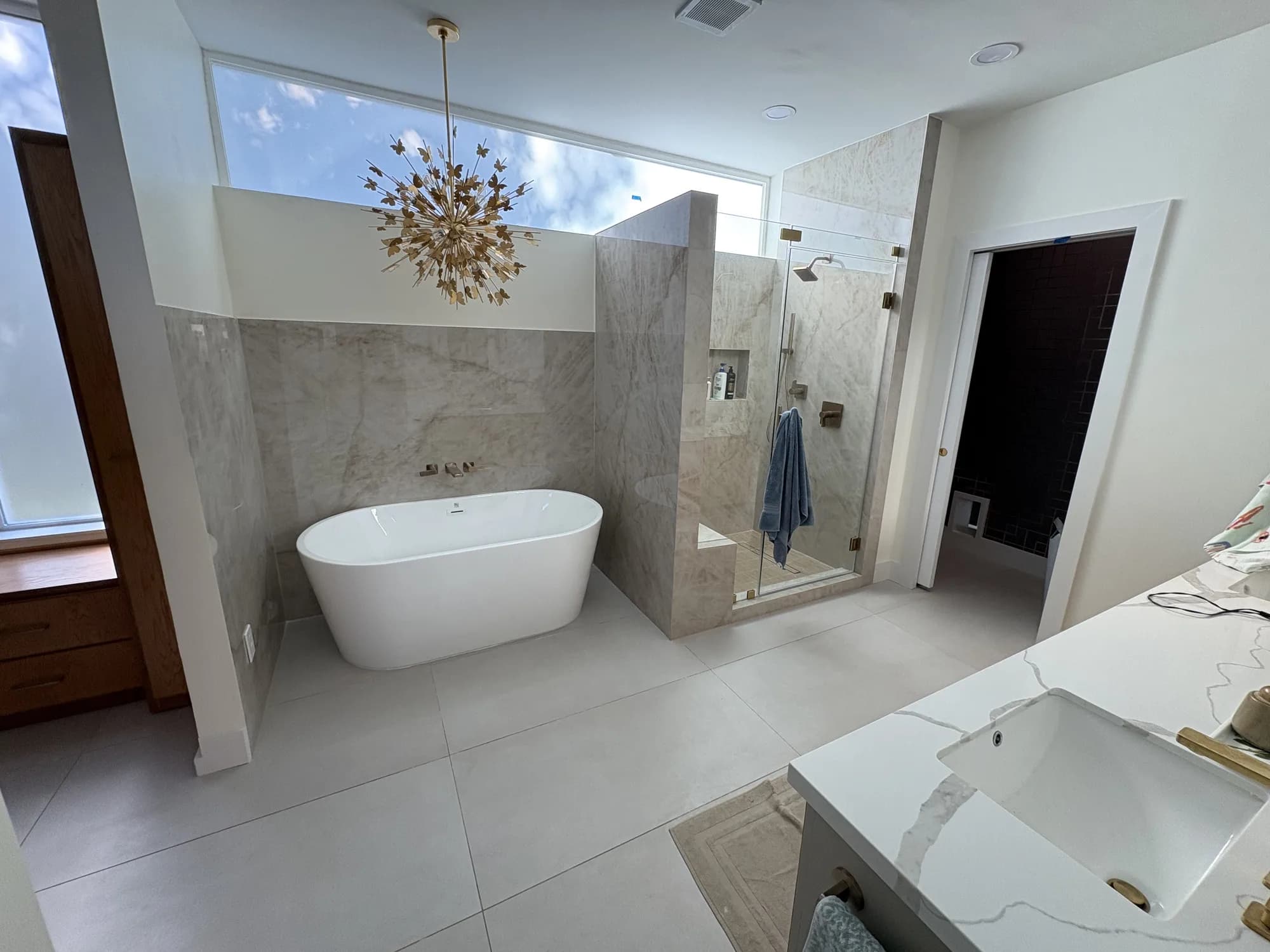 Wide view of a master bathroom with a freestanding tub, a marble walk-in shower with brass fixtures, a quartz-topped vanity, and a gold chandelier.