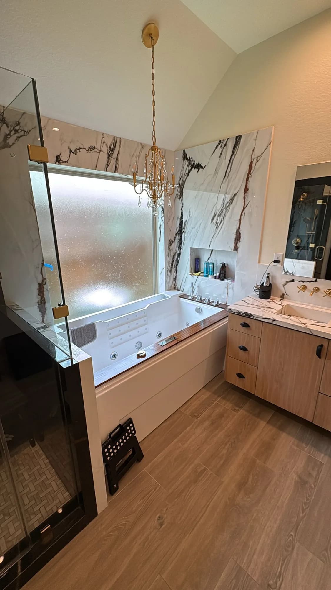 Bathroom with a dramatic white-and-burgundy veined marble accent wall behind a drop-in tub, a gold crystal chandelier, a frosted-glass window, a light-wood vanity with a marble waterfall counter and gold wall-mount faucet, and a black-glass walk-in shower.
