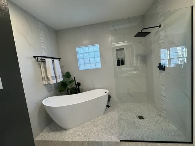 Bathroom with large-format white marble-look walls, a freestanding slipper tub on a pebble-mosaic platform, matte-black fixtures, a glass-block window, and a frameless glass walk-in shower with a black rain head.