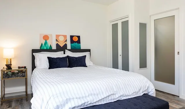 Bedroom in a converted-garage suite with an upholstered headboard, striped bedding with navy accent pillows, framed art prints, frosted-glass closet doors, and wood-look flooring.