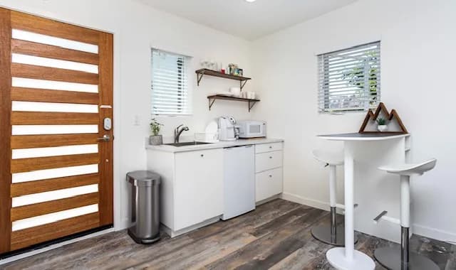 Converted-garage suite with a horizontal wood-slat entry door, a compact white kitchenette with open wood shelving and a bar sink, and a white pedestal bar-height table with stools on wood-look flooring.