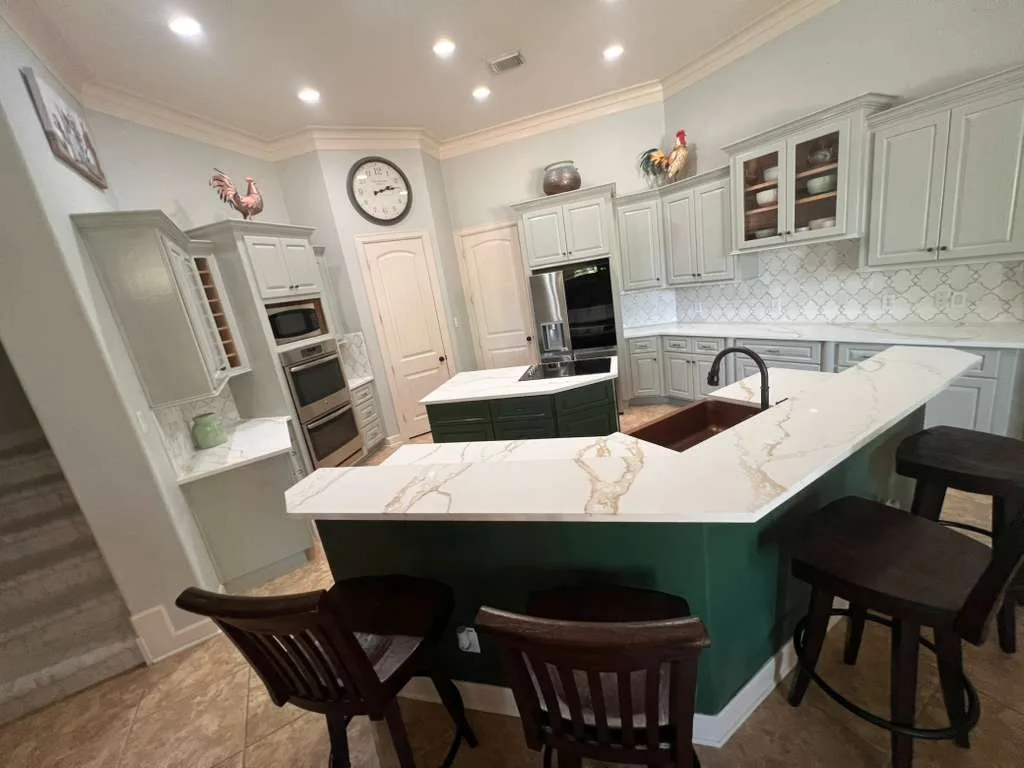 Sage-green kitchen island with a white quartz waterfall countertop, a copper farmhouse sink, a black gooseneck faucet, and wood bar stools, set against gray perimeter cabinets and an arabesque tile backsplash.