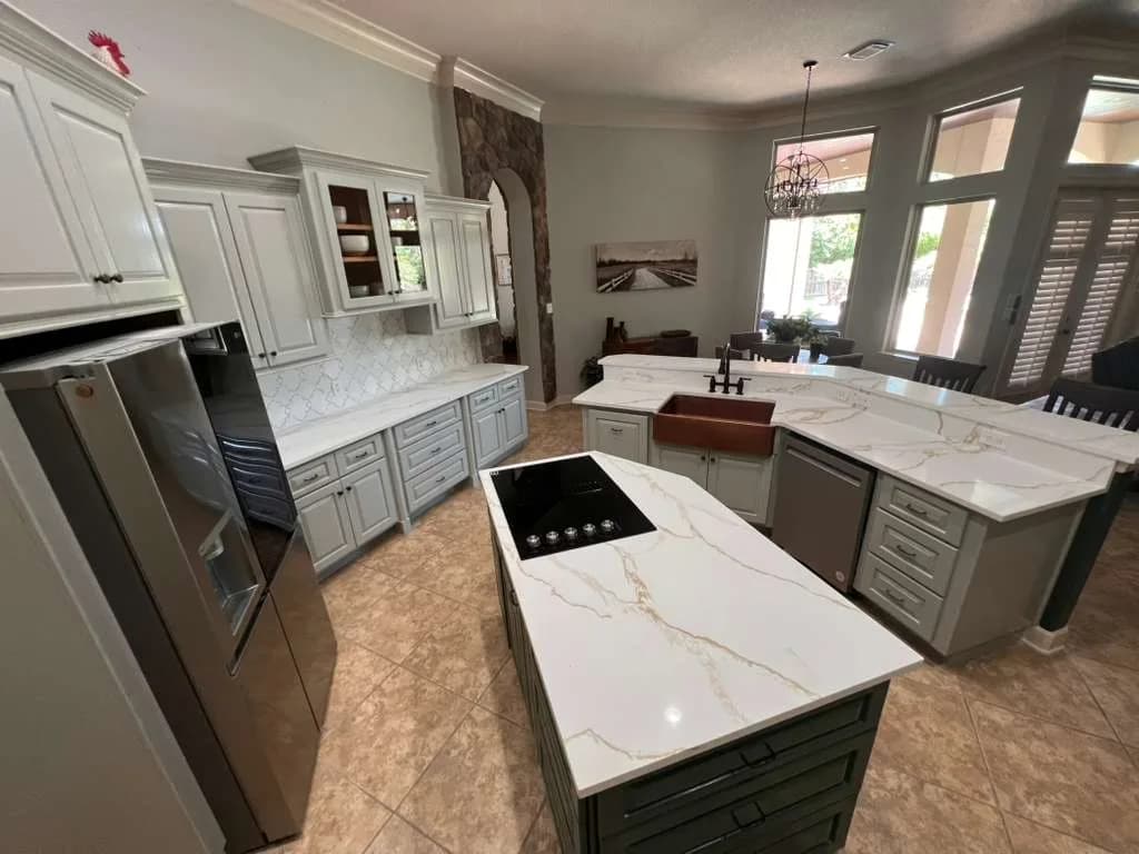 Open kitchen with a green cooktop island, white quartz counters with gold veining, gray cabinets, stainless appliances, and a stone-accent archway to the dining area.