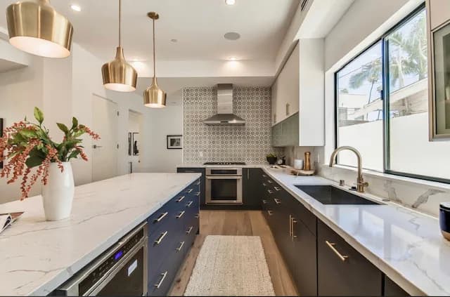 Kitchen with white upper cabinets and navy lower cabinets with gold pulls, white quartz countertops, a trio of brass dome pendants, and a stainless wall-mount hood over a patterned tile backsplash.