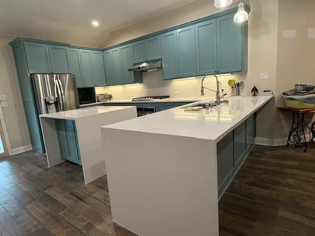 Teal shaker kitchen with a white quartz waterfall peninsula, dark wood floors, stainless appliances, glass pendant lights, and under-cabinet lighting.