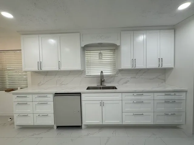 All-white shaker kitchen with white quartz countertops, a marble-look slab backsplash, an arched window niche with a pendant light, a stainless dishwasher, and marble-look tile flooring.