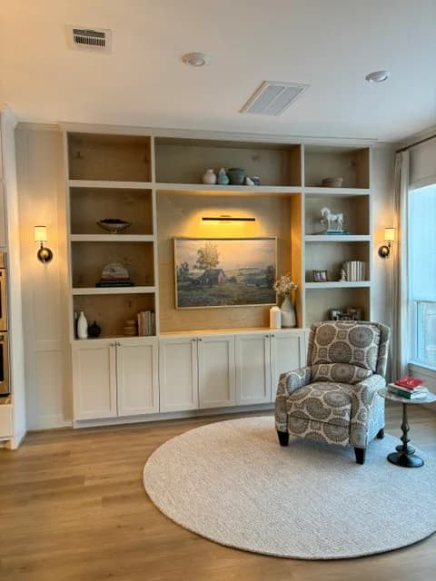 Living room with a full-wall custom built-in of white cabinetry and wood-backed open shelving, framed landscape art lit by a picture light, and a patterned accent chair on a round rug.