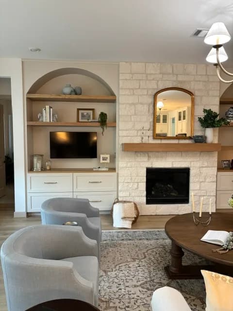 Living room with a white stone fireplace and wood mantel, an arched built-in niche holding a TV and floating wood shelves, and gray upholstered swivel chairs around a round wood coffee table.