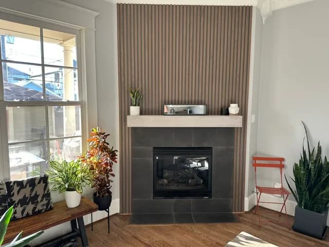 Living room with a floor-to-ceiling vertical wood-slat fireplace surround, a gray large-format tile hearth, a light-wood floating mantel, a gas fireplace, and a bright window with a front-porch view.