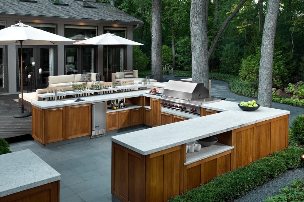 Custom outdoor kitchen with a built-in grill, stone countertops, and covered patio seating in a wooded backyard.