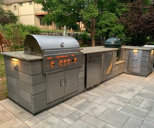 Backyard outdoor kitchen with a stainless built-in grill, a ceramic kamado cooker, stone-veneer cabinetry with granite countertops, and a paver patio.