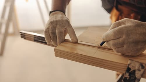 A carpenter in work gloves marking a measurement on a wood board at a job site.